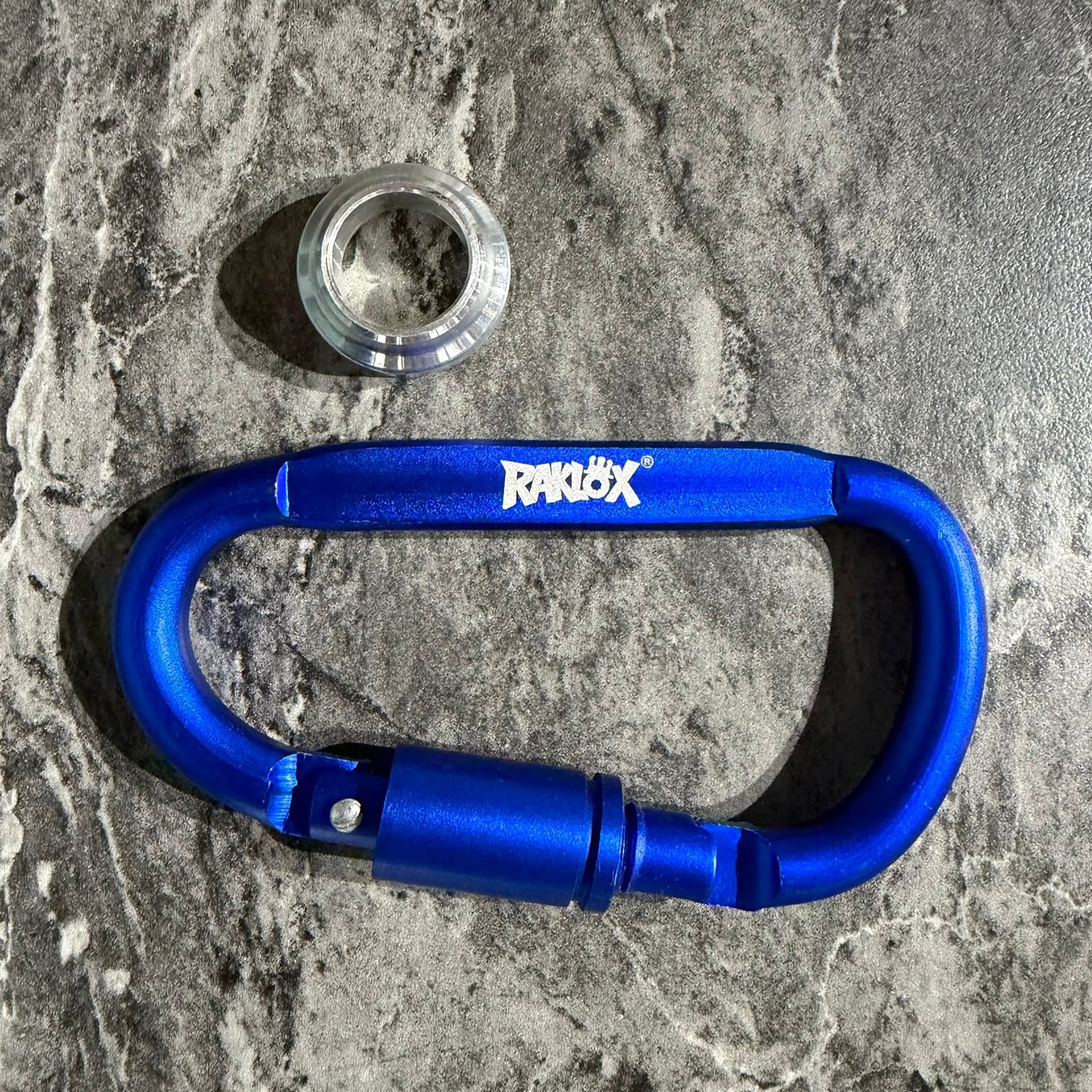 A bright blue anodized aluminum carabiner with a white RAKLOX logo on the side, displayed on a gray marbled surface.