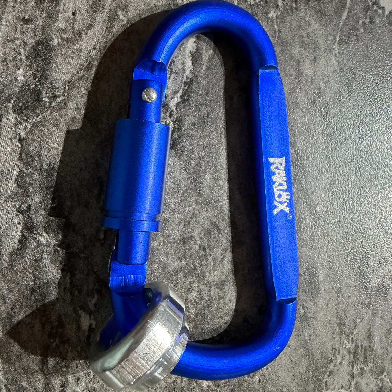A bright blue anodized aluminum carabiner with a white RAKLOX logo on the side, displayed on a gray marbled surface.