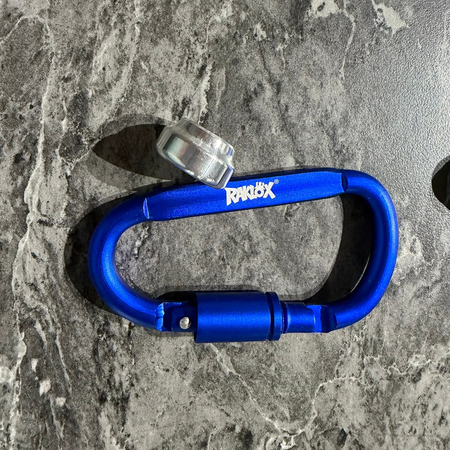 A bright blue anodized aluminum carabiner with a silver metal lock and a white RAKLOX logo on the side, resting on a gray marble-patterned surface.